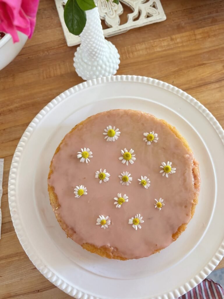 Pink glazed homemade cake topped with small fresh daisies on a white cake stand for a 2nd birthday