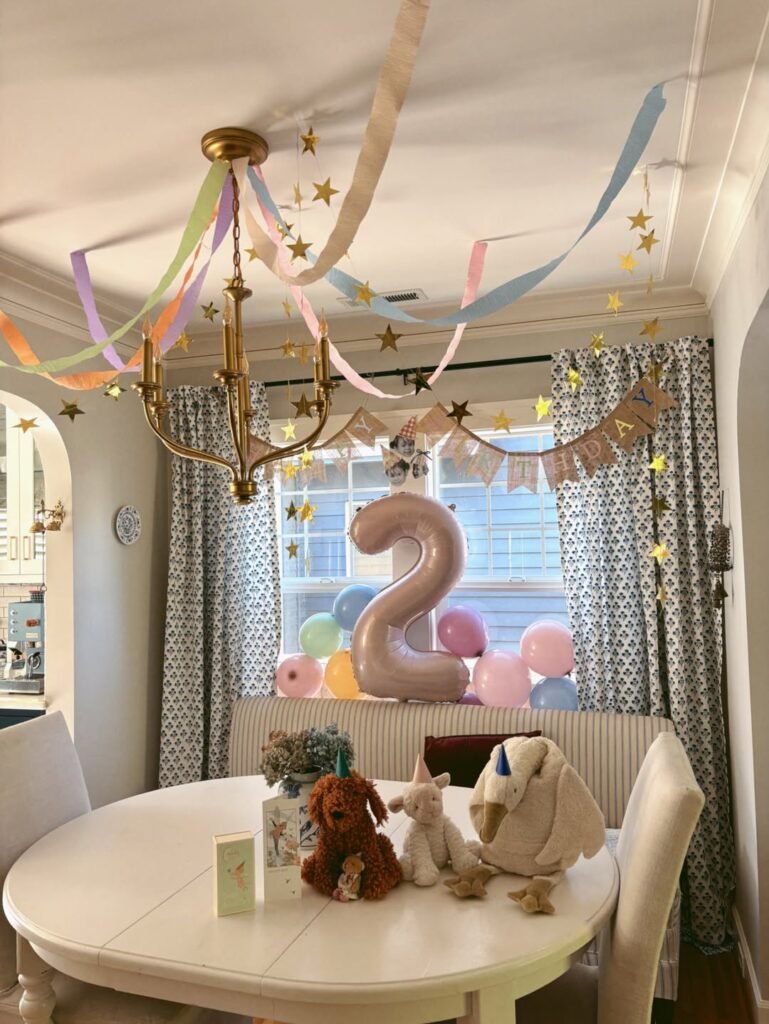Cozy dining room set up for a pastel 2nd birthday with streamers, gold stars, a 2 balloon and plush toys on the table