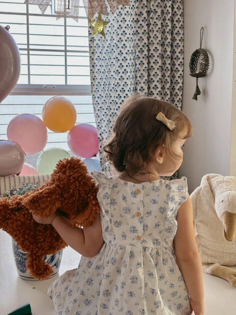Back view of a birthday girl in a floral dress with bow hair clips holding a teddy bear