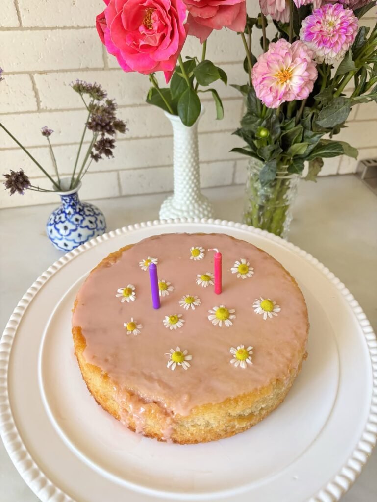 Pink daisy birthday cake with pink and purple candles next to a vase of pink roses and dried flowers
