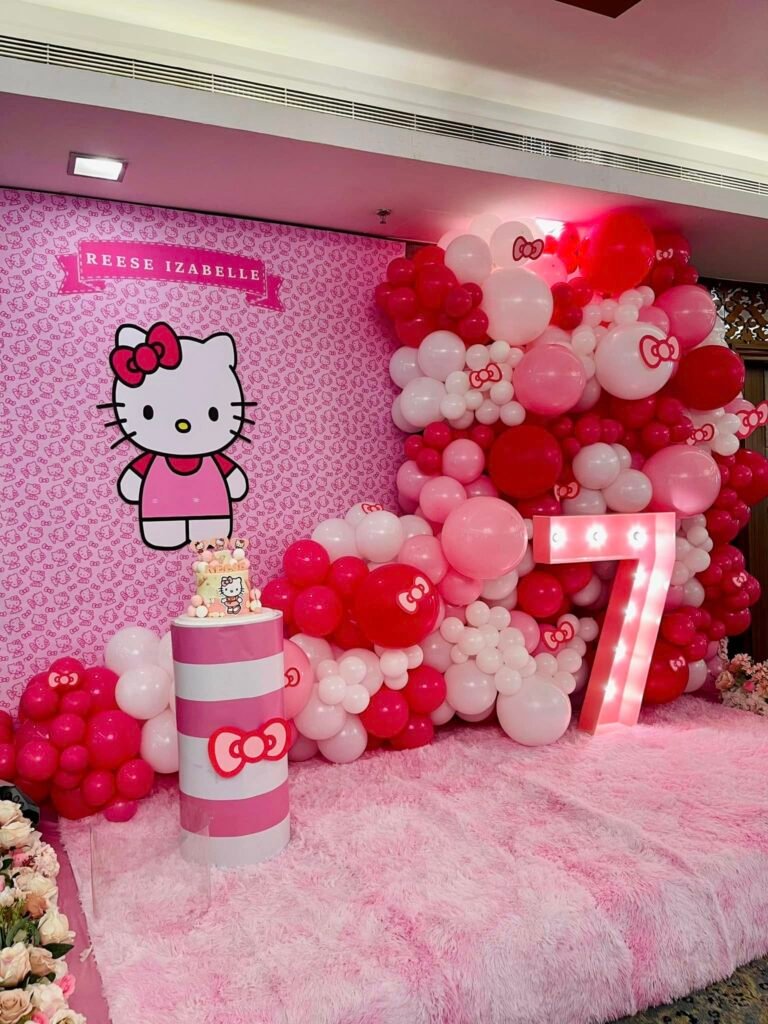 Hello Kitty backdrop with a light up number 7, pink and red balloons, a cake on a pink stand and a pink fur rug