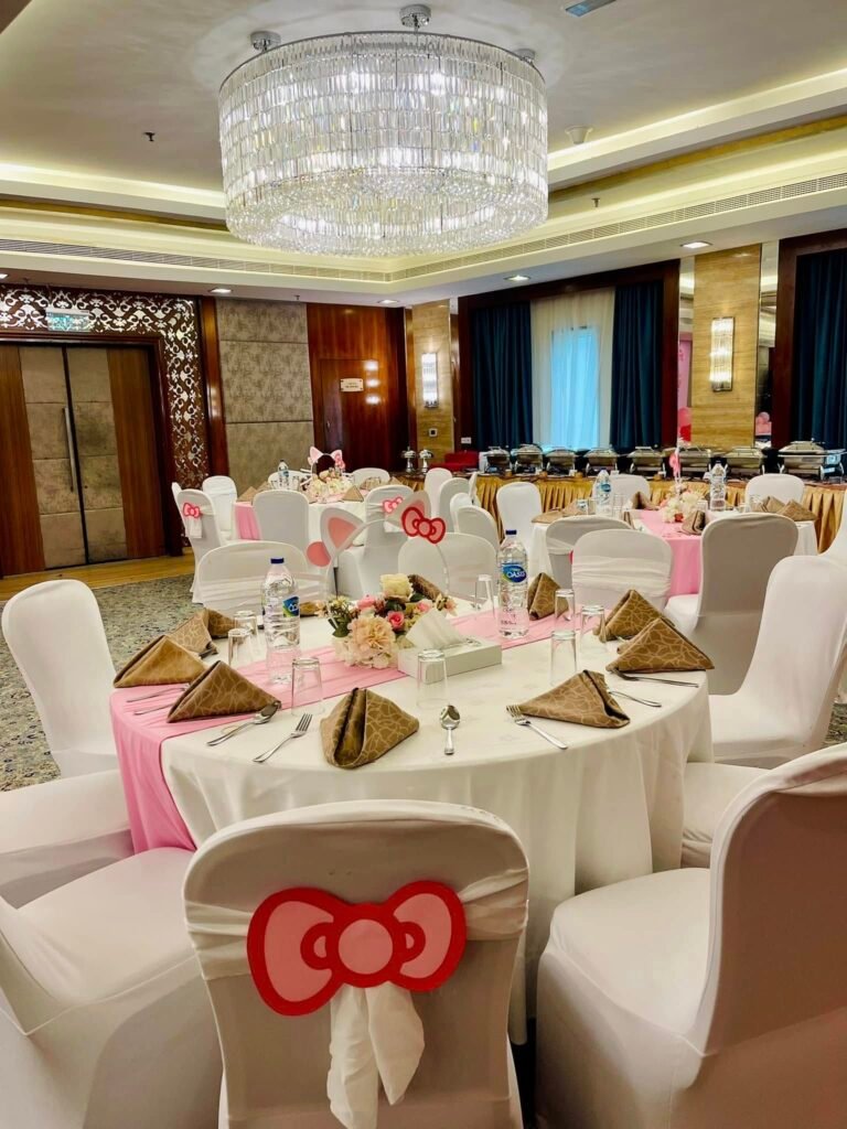 Ballroom party tables with white linens, pink napkins and white chair covers tied with red Hello Kitty bows