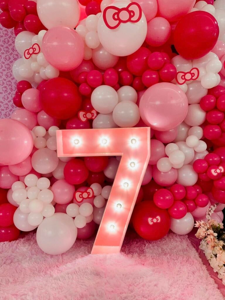 Light up marquee number 7 in front of a pink, red and white balloon wall with bow balloons