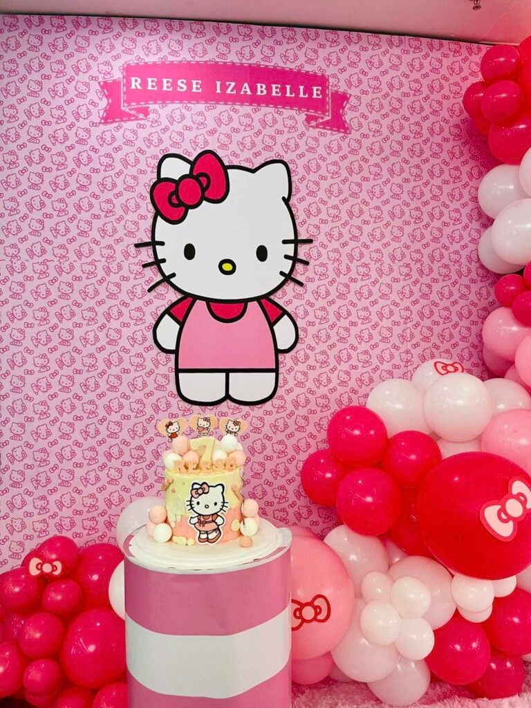Large Hello Kitty cutout on a pink Hello Kitty print wall with a birthday cake on a pink and white stand