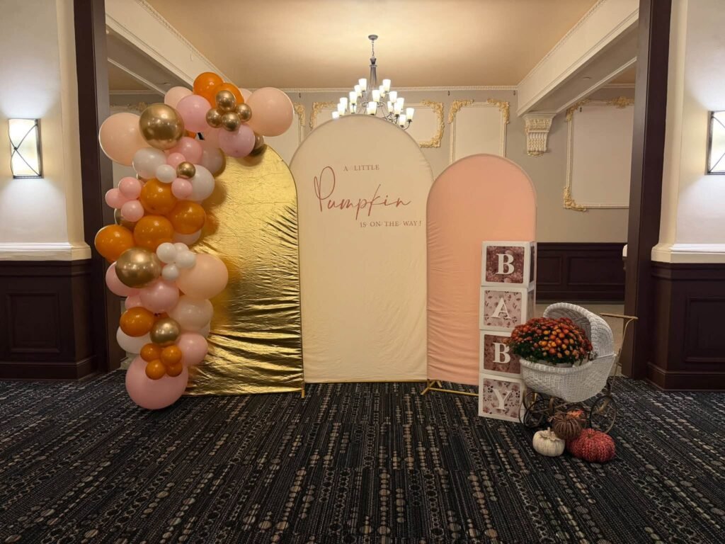 Little Pumpkin baby shower backdrop with gold, cream and peach arch panels, a pink, orange and gold balloon garland and BABY blocks