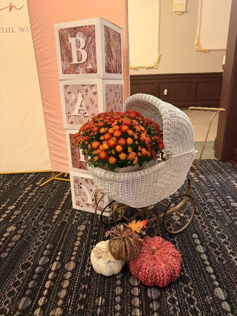 BABY letter blocks next to a white wicker baby carriage filled with orange mums and small pumpkins on the floor