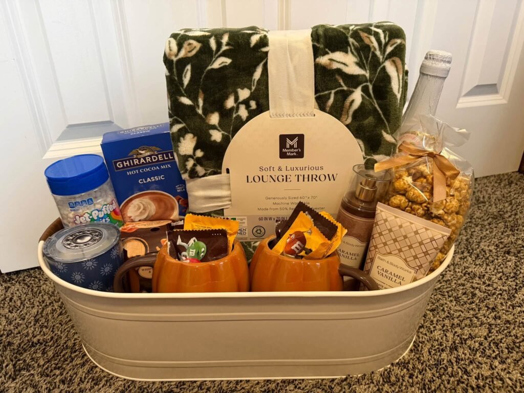 Fall gift basket in a white metal tub with a lounge throw, pumpkin mugs, hot cocoa, Ghirardelli chocolate and popcorn