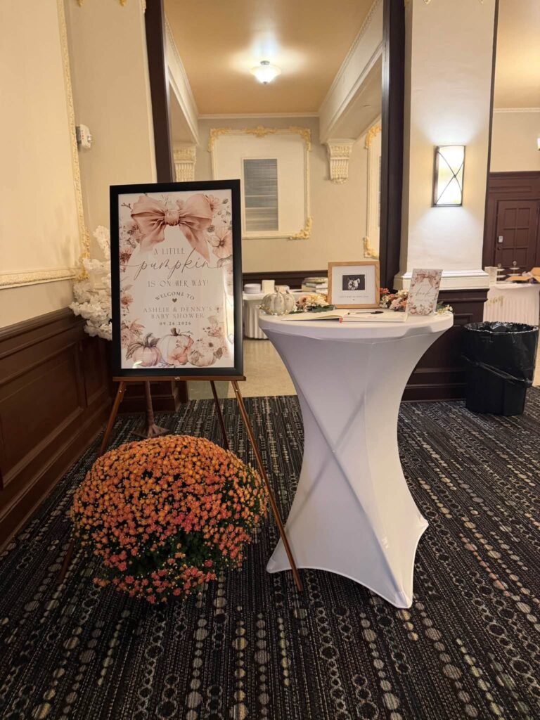 Pink floral Little Pumpkin welcome sign with a big bow on an easel, a pot of orange mums and a cocktail table with a sonogram frame