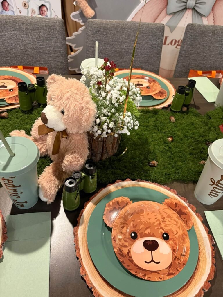 Teddy bear sitting on a moss table runner next to bear face plates and baby breath