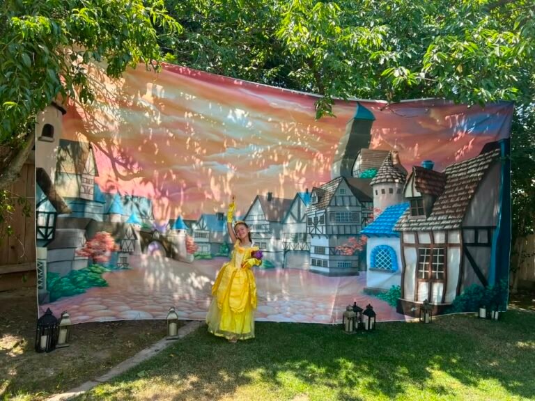 Birthday girl in a yellow Belle dress in front of a village backdrop in the yard