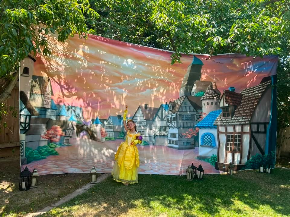 Birthday girl in a yellow Belle dress in front of a village backdrop in the yard
