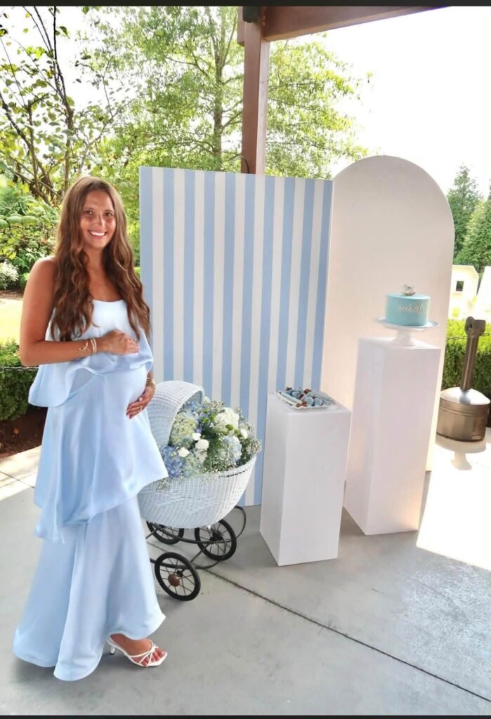 Mom to be holding her dress by a blue striped arch backdrop and pram