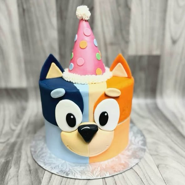 Bluey and Bingo face birthday cake, half blue and half orange, with a pink party hat on top