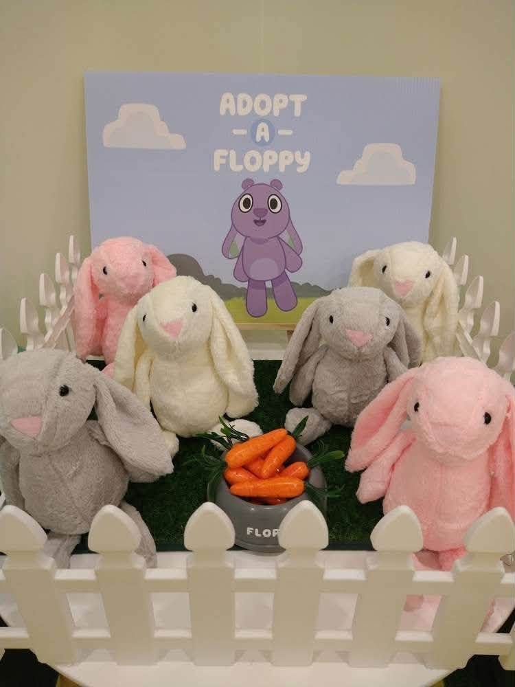 Adopt a Floppy station with plush bunnies inside a white picket fence on grass with a bowl of carrots