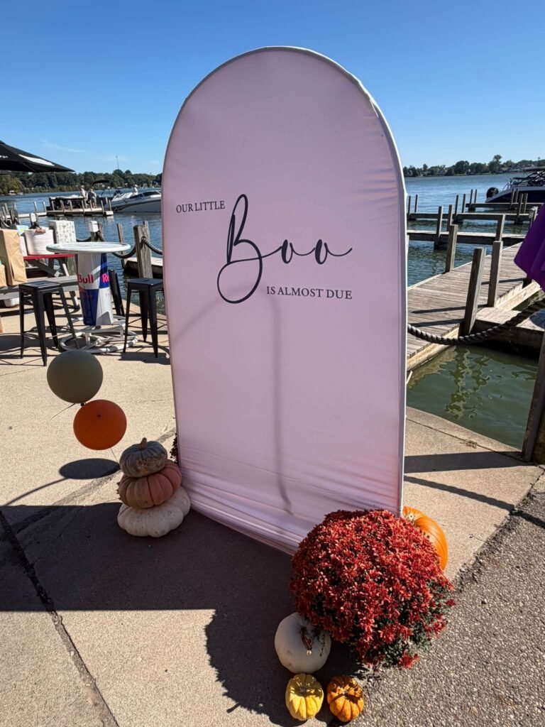 Little Boo baby shower arch sign that says Our Little Boo is almost due with pumpkins, red mums and balloons by a lake