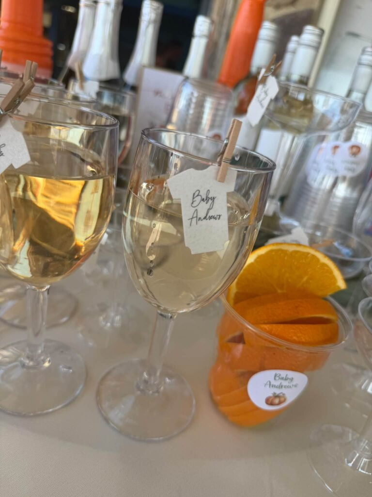 Wine glasses with tiny baby bodysuit tags clipped on with clothespins and orange slices at a baby shower drink station