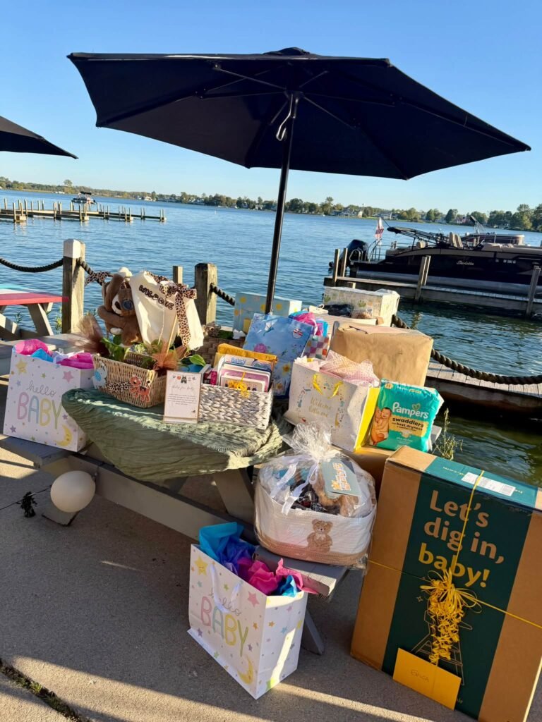 Outdoor baby shower gift table by a lake under an umbrella with gift bags, diapers and presents