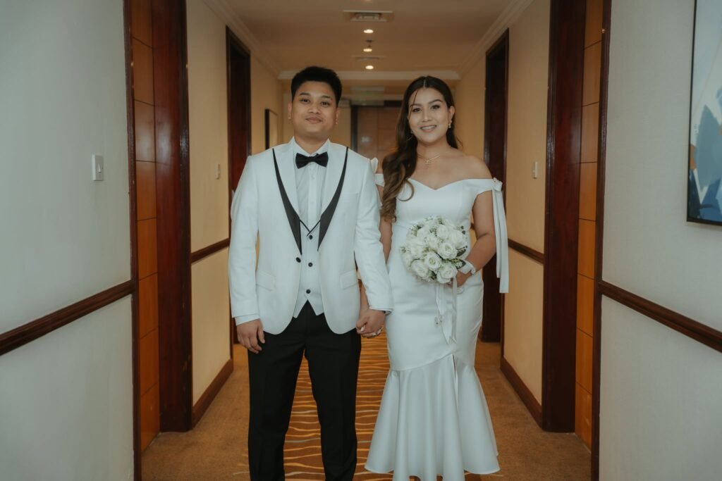 Groom in a white tuxedo jacket and bride in a white mermaid dress walking down a hotel hallway