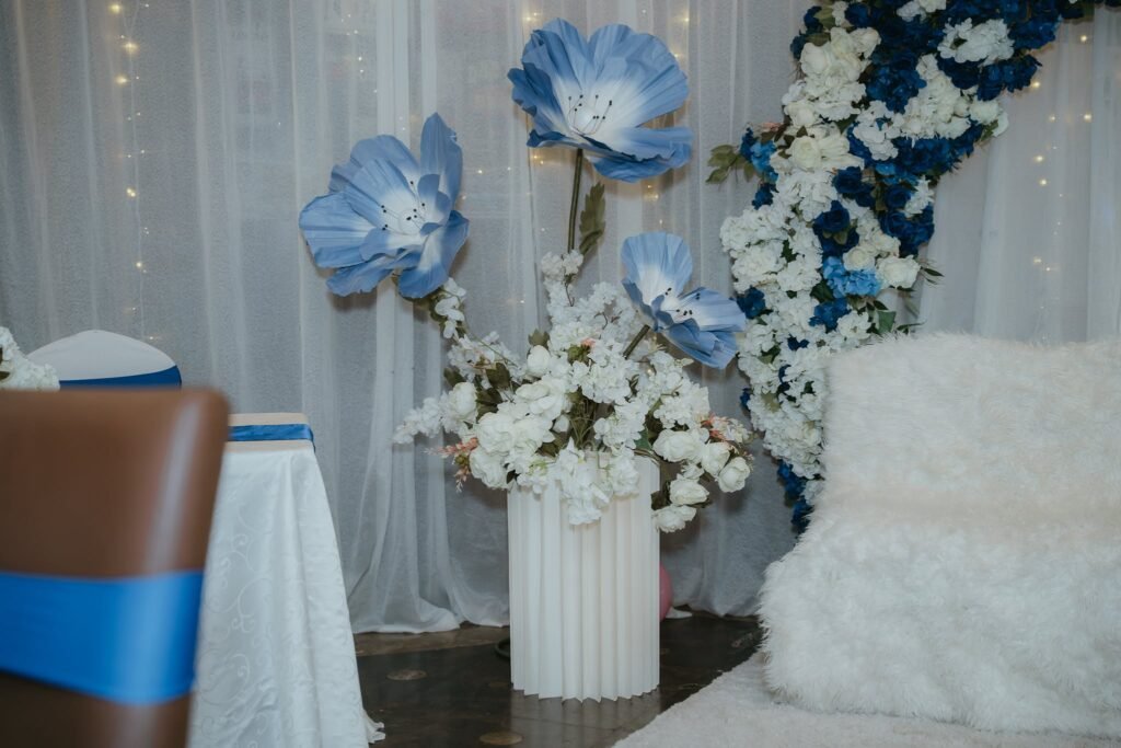 Giant blue paper flowers and white blossoms in a white column vase next to a fluffy chair