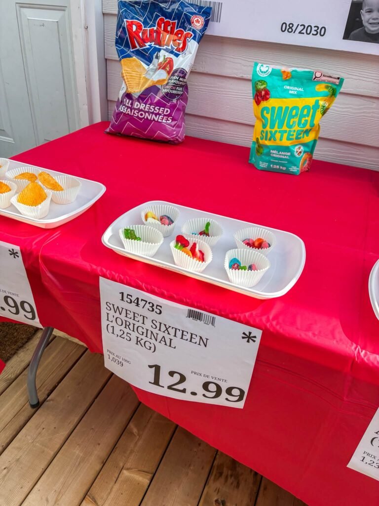 Snack table with store style price tag signs and sample cups on a red tablecloth