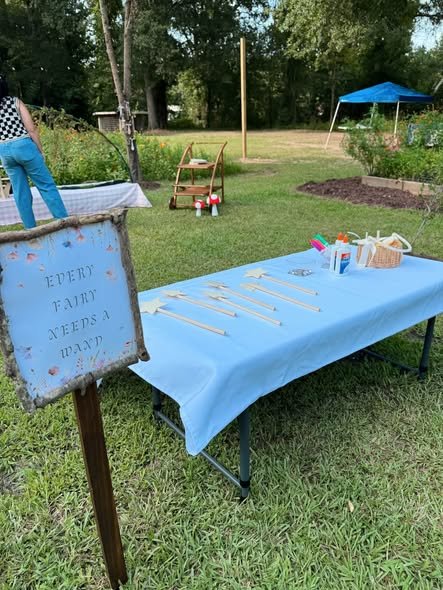 Every Fairy Needs a Wand sign on a stake next to a blue table with wand making supplies in a garden