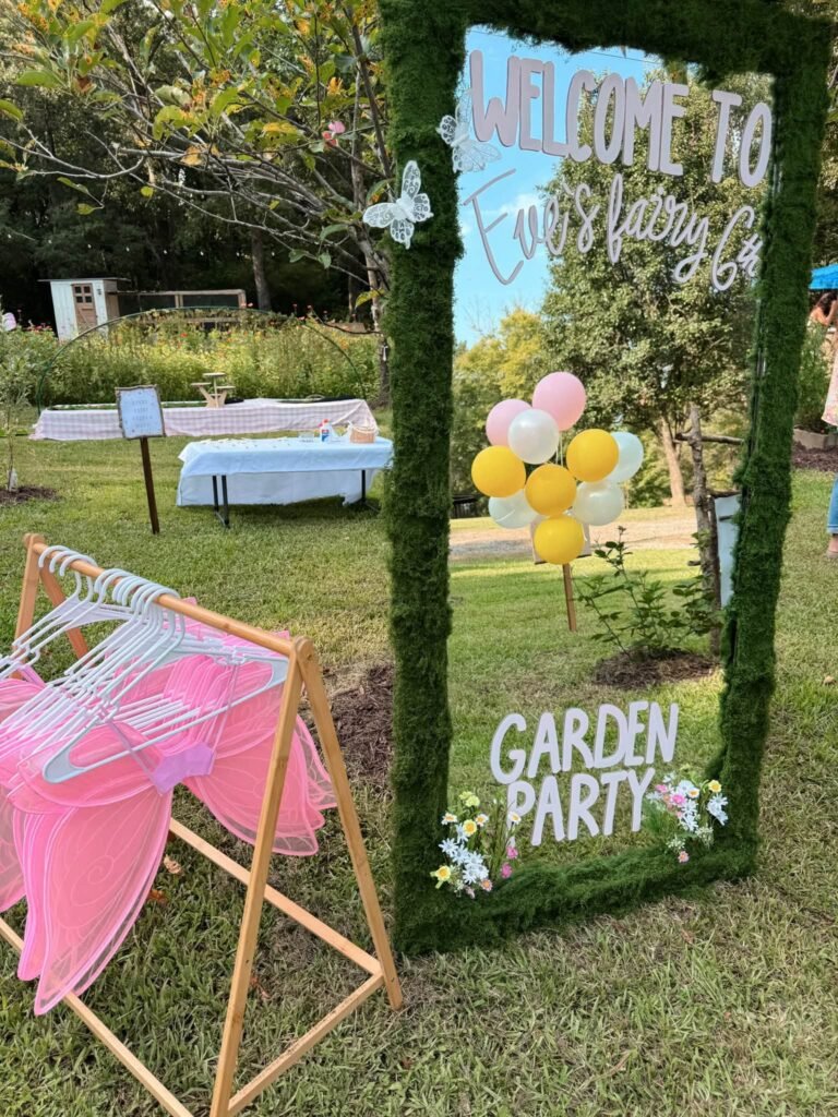 Moss framed welcome mirror sign for a fairy garden birthday party next to a wooden rack of pink fairy wings and yellow balloons