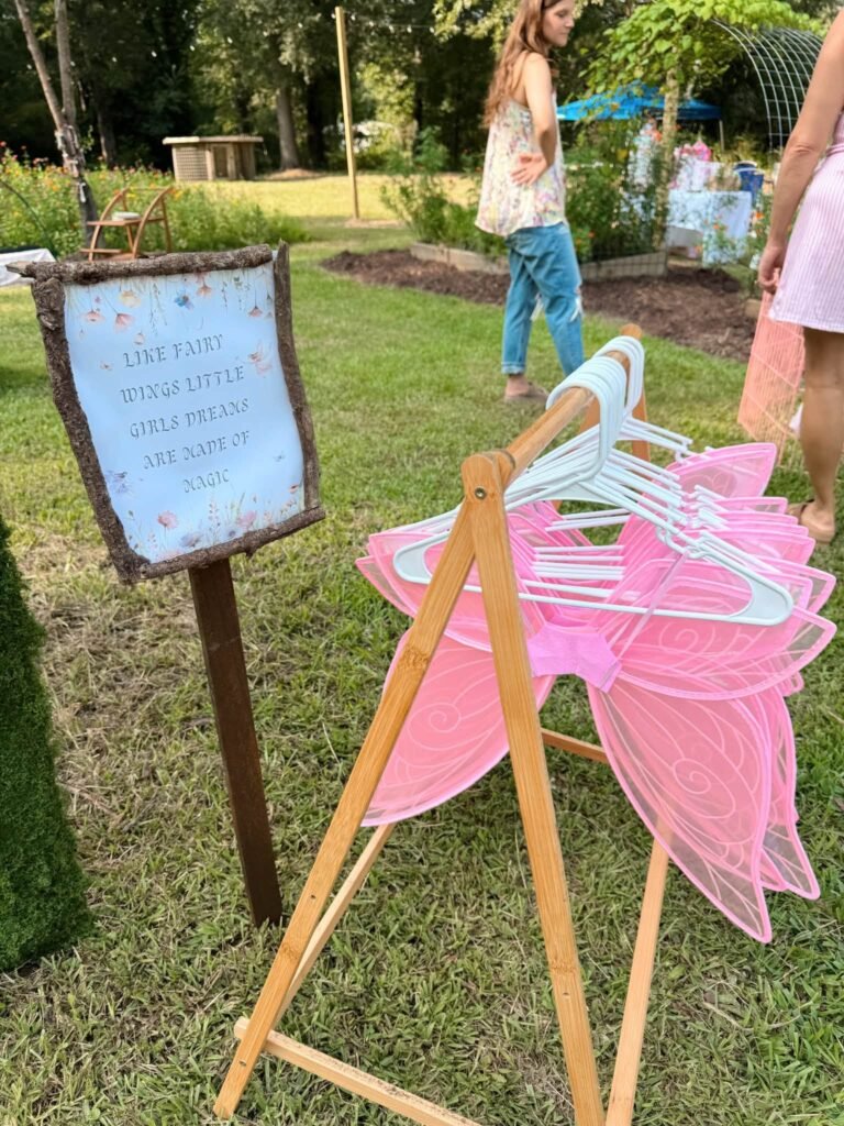 Wooden rack of pink fairy wings on hangers next to a framed fairy sign on a stake in a garden