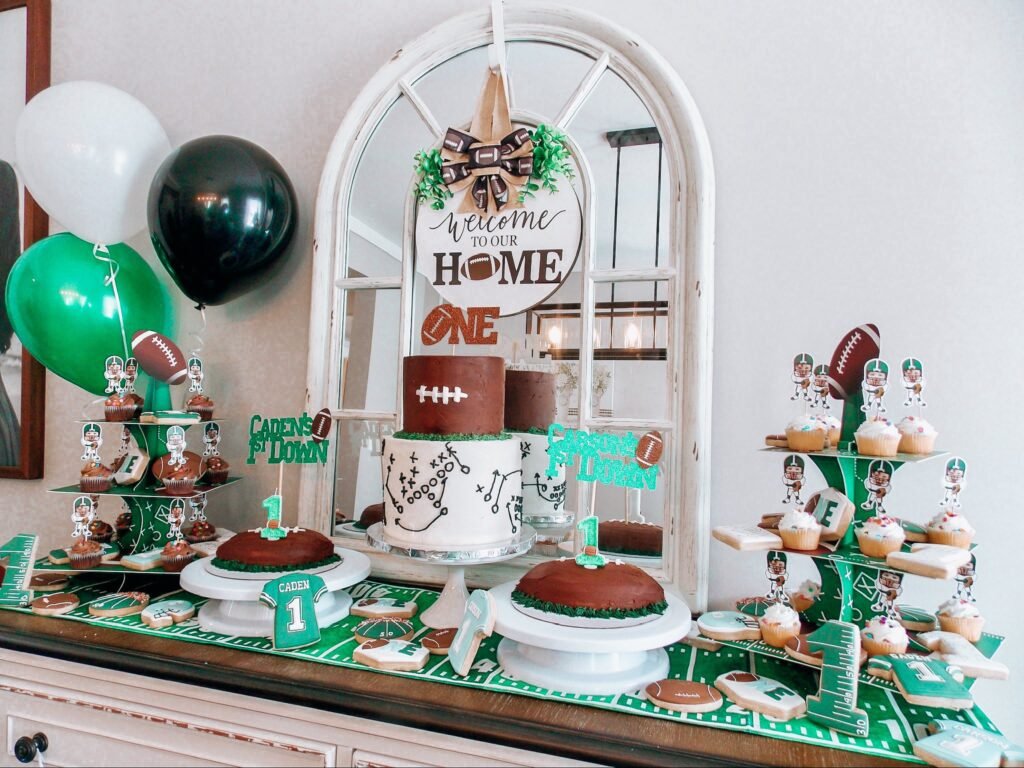 Football first birthday dessert table with a Welcome to the Home of ONE sign, green and black balloons and football cakes