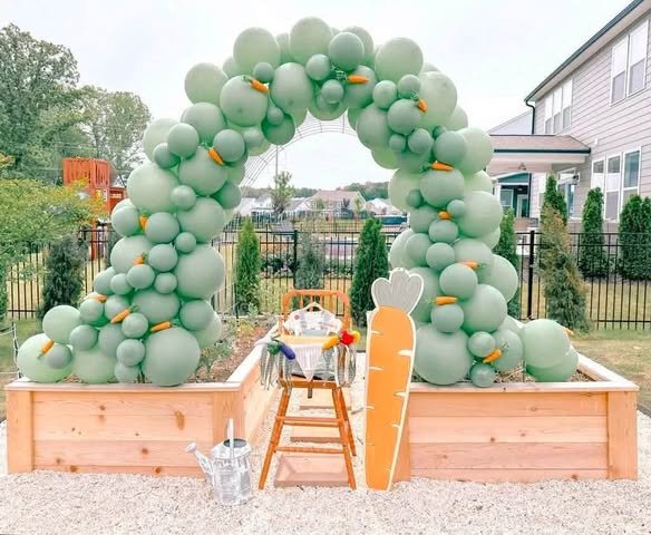 Sage green balloon arch over a raised wooden garden bed with a carrot cutout and a high chair outdoors