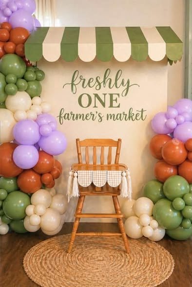 Freshly One farmers market 1st birthday backdrop with a green striped awning, a wooden high chair and green, lilac and rust balloons