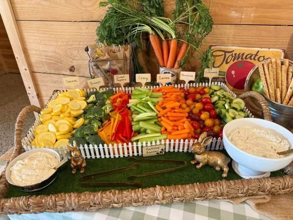 Veggie tray garden with rows of carrots, peppers, cucumbers and dips on faux grass with a white picket fence