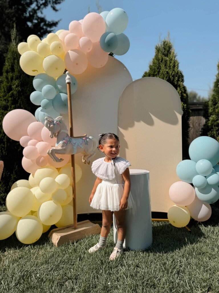 Birthday girl in a white dress in front of a pastel balloon arch