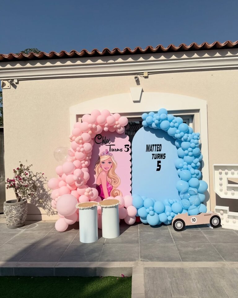Outdoor twins birthday setup with pink Barbie and blue race car balloon arches, white plinths and a race car cutout