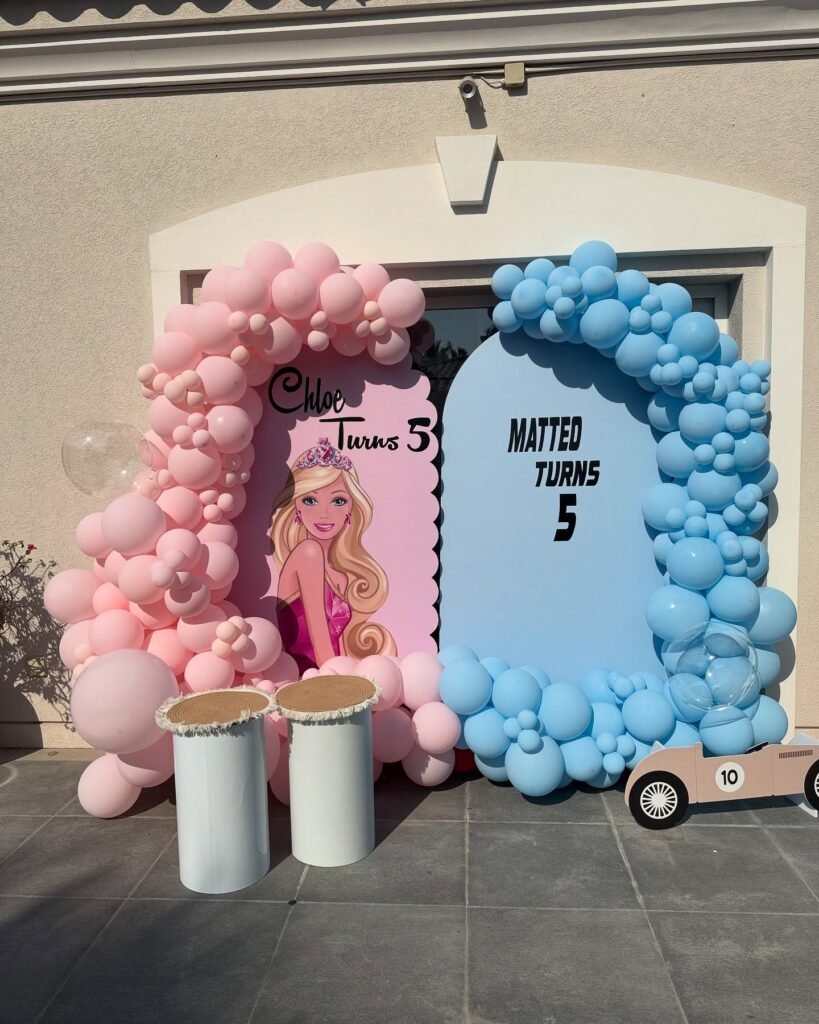 Twins 5th birthday backdrop with a pink Barbie arch and a blue race car arch framed by pink and blue balloon garlands