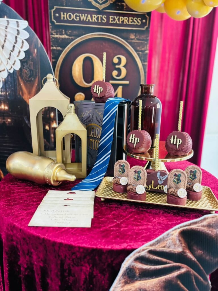 Harry Potter baby shower dessert display with a Platform 9 3/4 backdrop, gold lanterns, chocolate covered apples and house crest treats