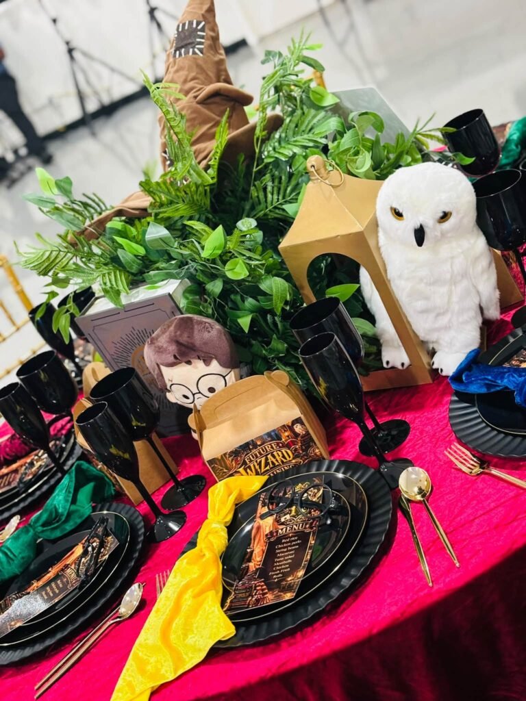 Harry Potter baby shower centerpiece with greenery, a sorting hat, a plush snowy owl, gold lanterns and black goblets