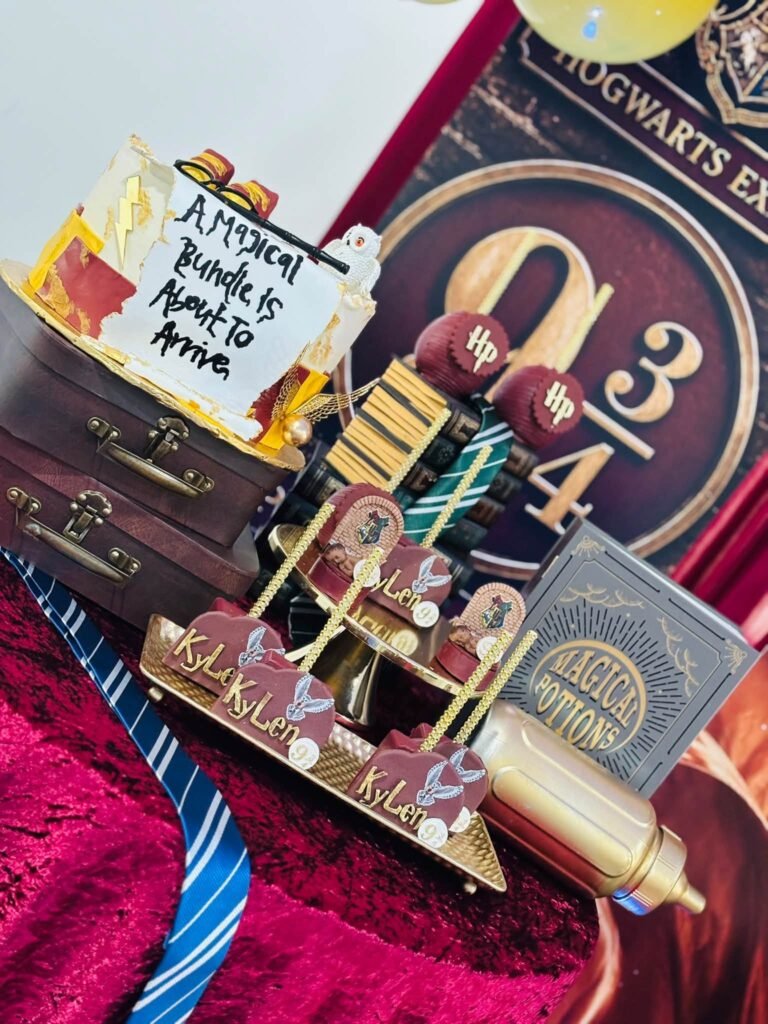Side view of the Magical Bundle baby shower cake on suitcases next to cake pops and chocolate apples in front of a Platform 9 3/4 backdrop