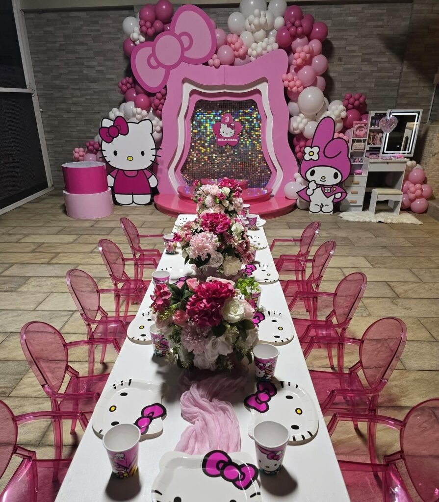 Long white table with pink ghost chairs, Hello Kitty plates and pink flower centerpieces in front of the backdrop