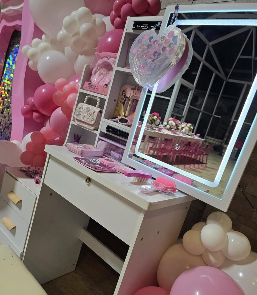 Glam vanity station with a white desk, a light up mirror and pink accessories next to pink balloons