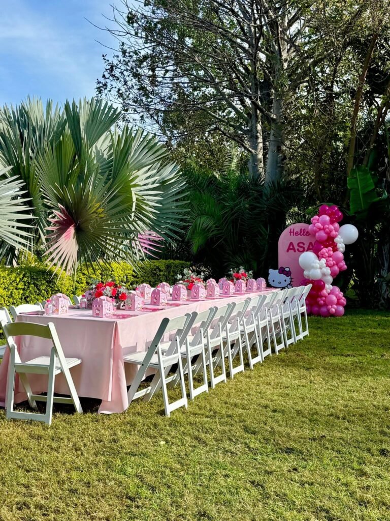 Long pink party table with white chairs set on a garden lawn with a balloon sign at the end