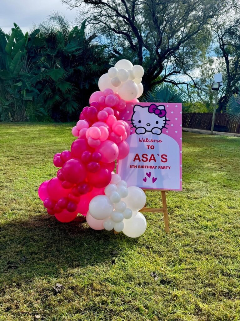 Hello Kitty welcome sign on an easel next to a pink and white balloon column on a lawn