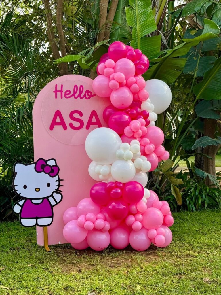 Pink Hello name sign with a Hello Kitty cutout and a pink and white balloon garland in a garden