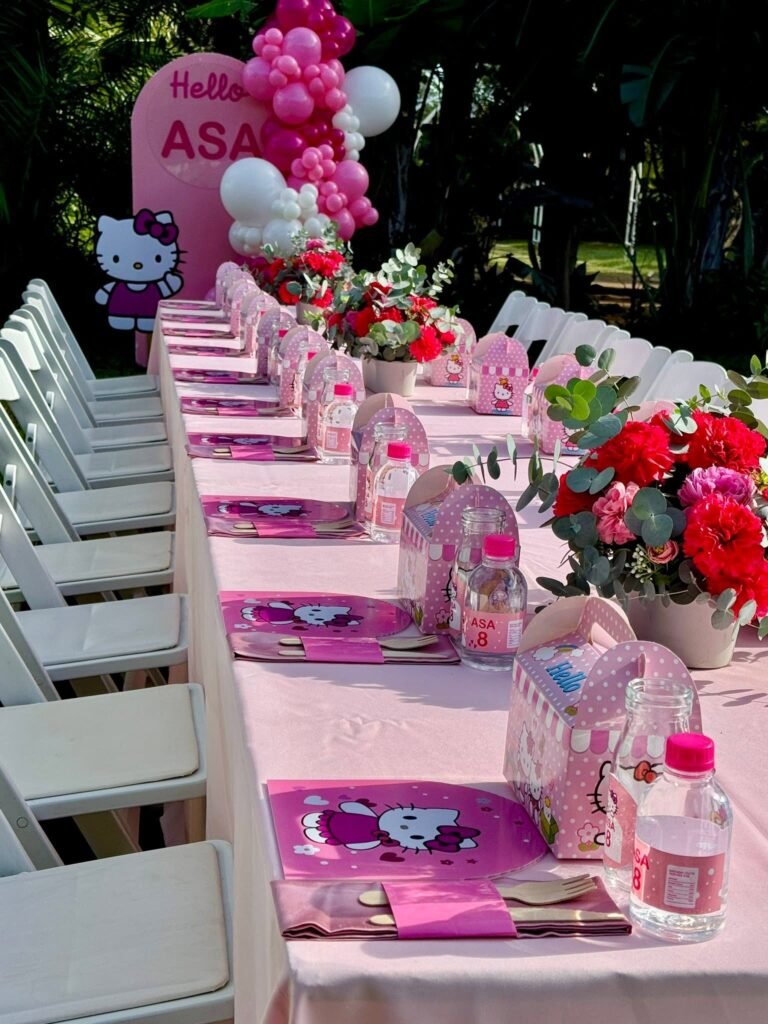 Long Hello Kitty party table with pink cloth, red roses and white chairs in a garden