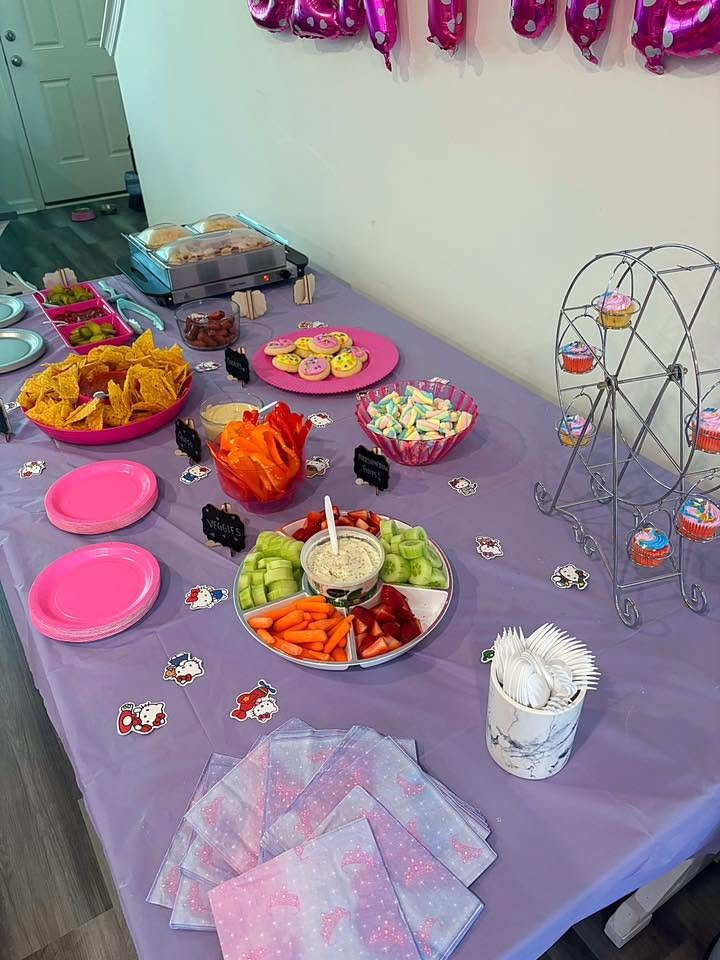 Hello Kitty party snack table with chips, a veggie tray with dip, fruit, pink plates and a Ferris wheel cupcake stand