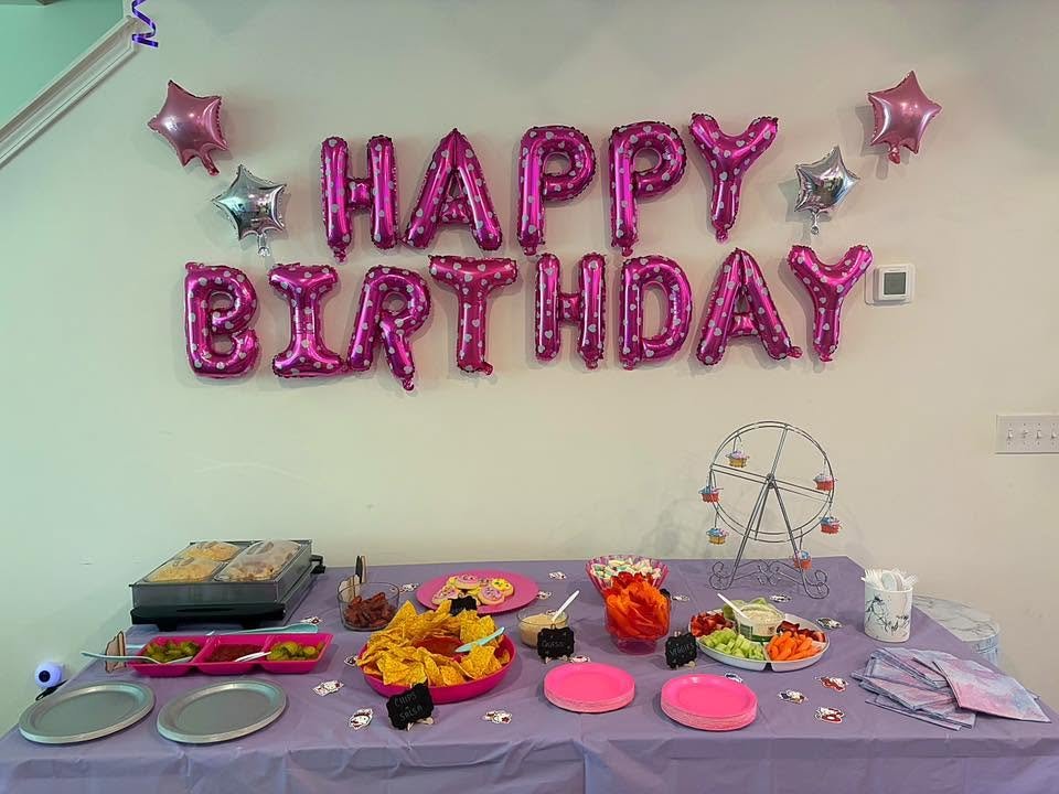 Pink foil Happy Birthday balloon letters above a lilac snack table with chips, dips and a Ferris wheel stand