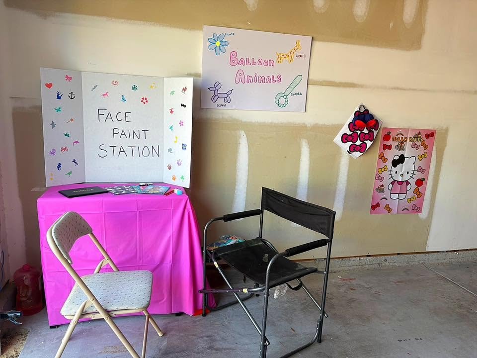 Face paint station with a hand drawn sign, a pink table, a folding chair and a Hello Kitty poster