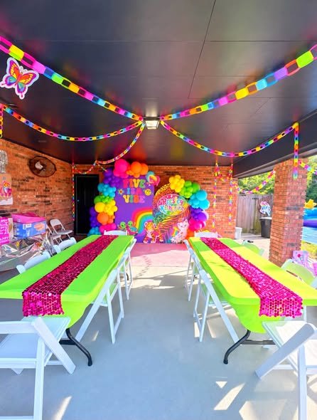 Lisa Frank birthday patio with neon pink and lime table runners, rainbow streamers and a rainbow balloon backdrop