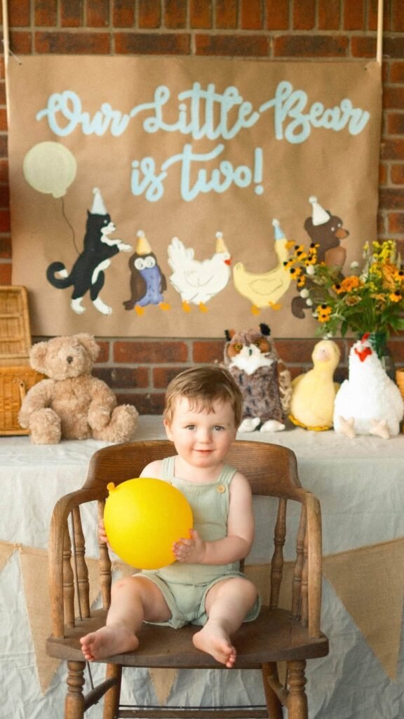 Birthday boy in a sage green romper on a wood chair holding a yellow balloon