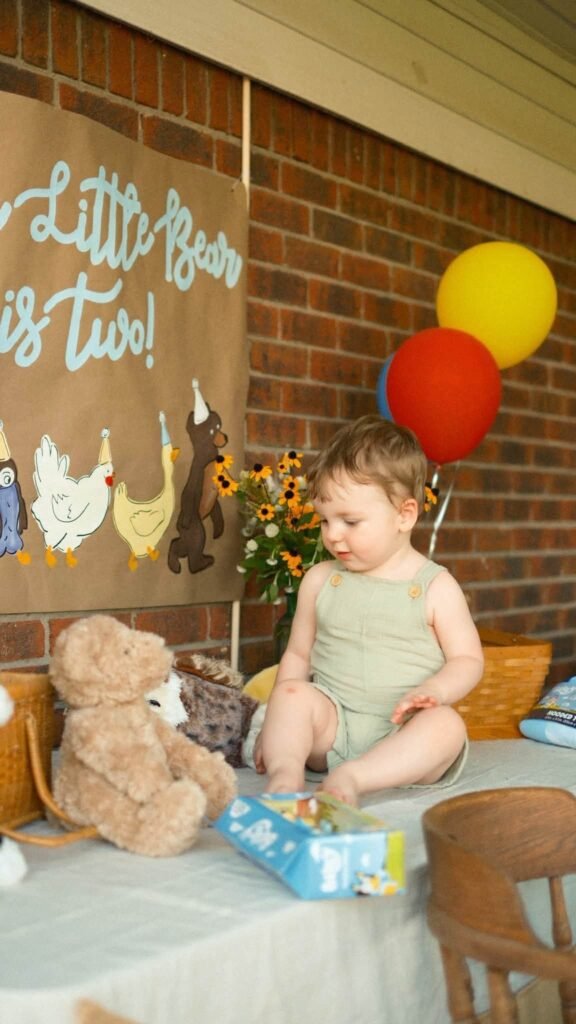 Birthday boy in a sage romper sitting on the party table next to a teddy bear