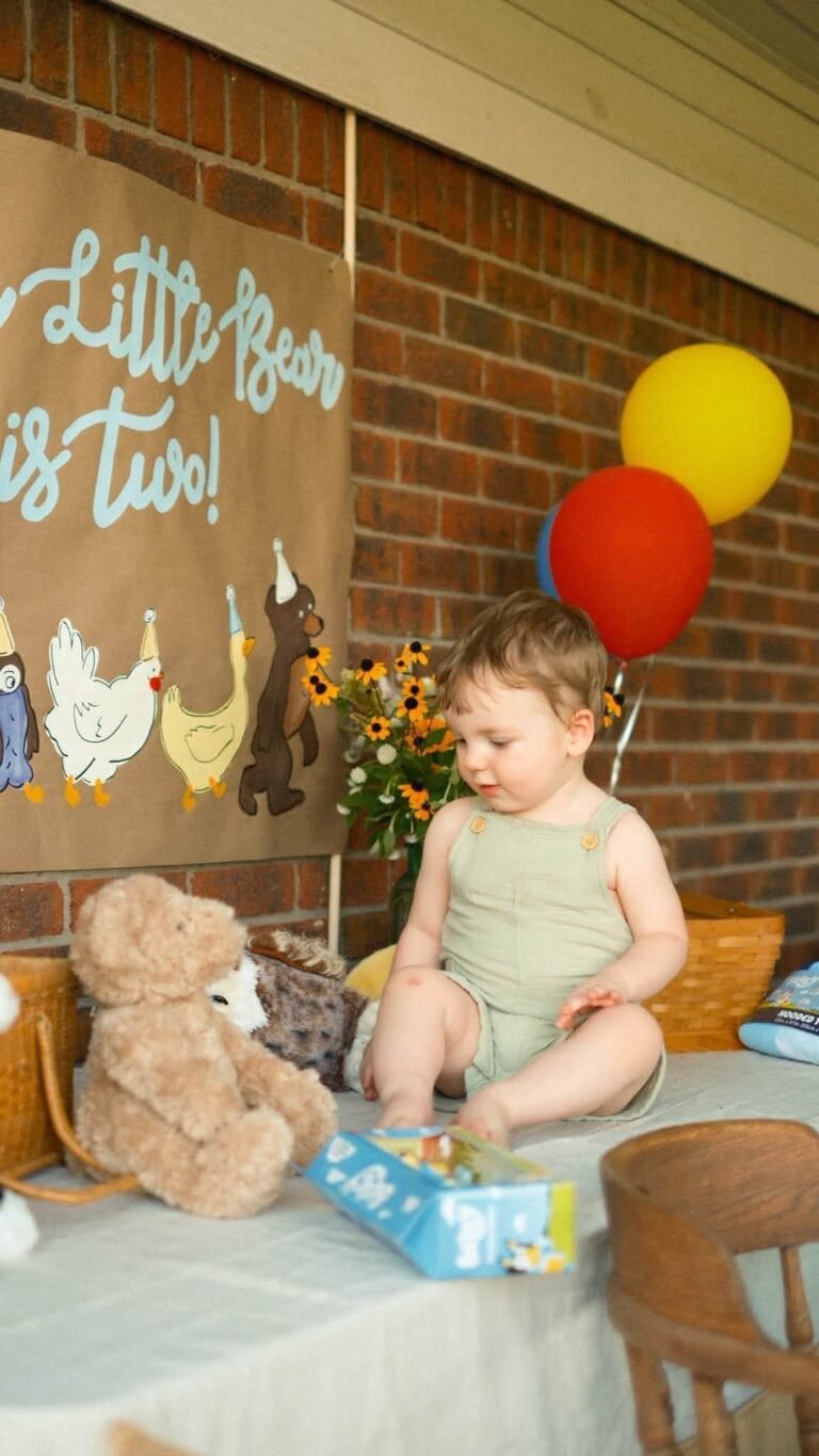 Birthday boy in a sage romper sitting on the party table next to a teddy bear